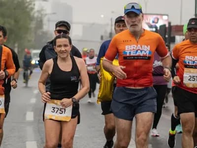 Un grupo de corredores avanza por una avenida durante una maratón en un día nublado; varios llevan polera del Brain Team y el número de competencia en el pecho.